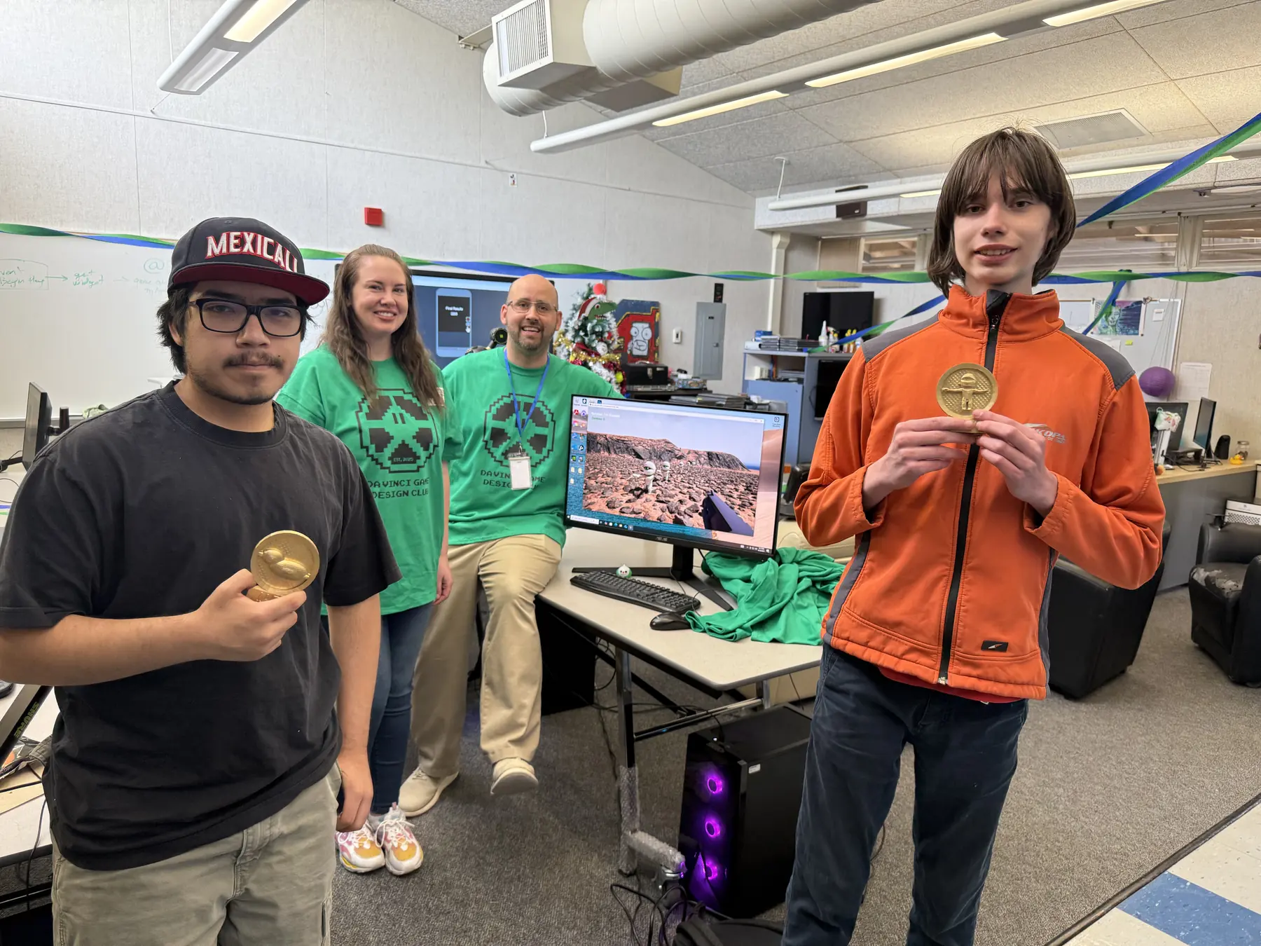Two students holding gold club medals in the Da Vinci game lab, with instructors behind them